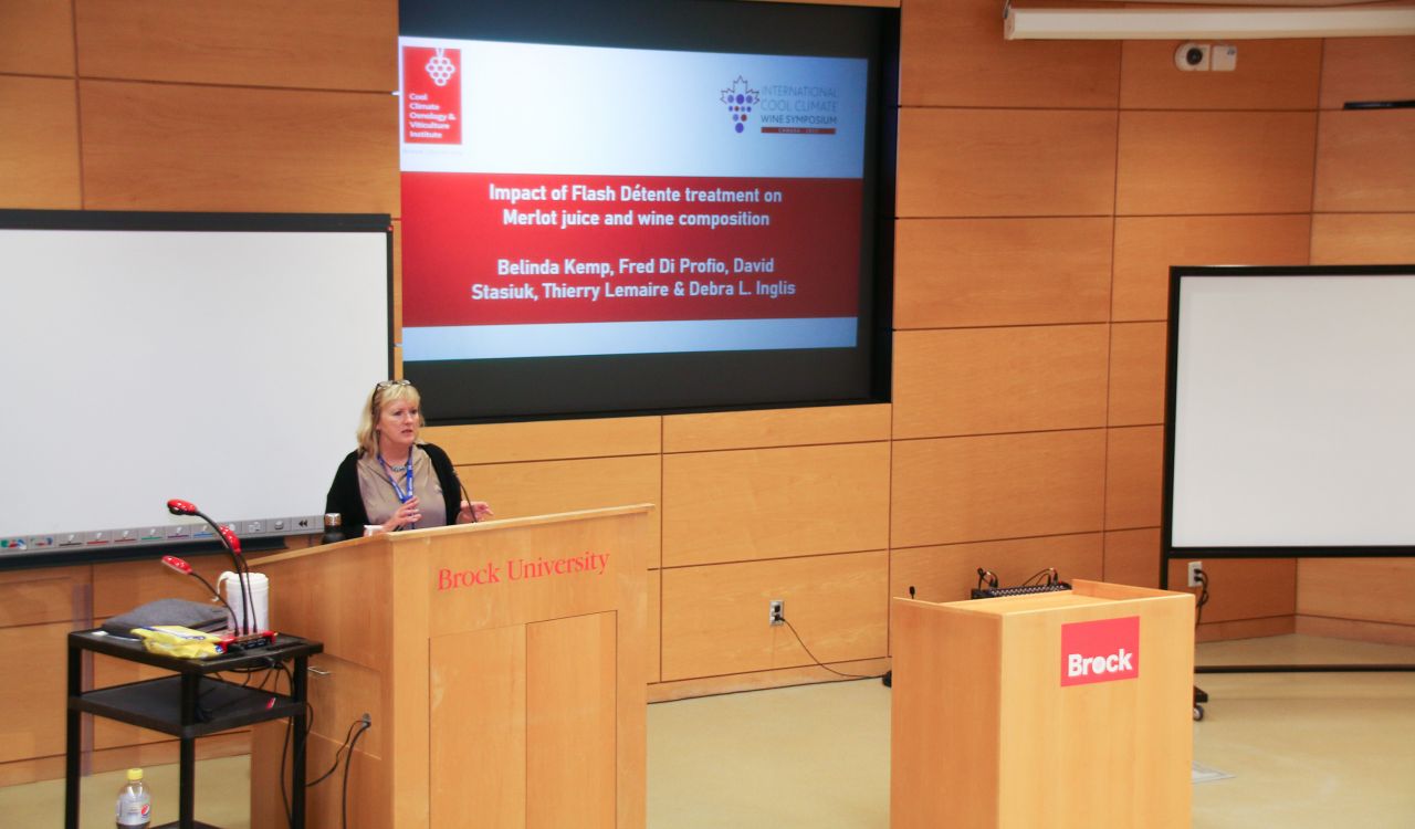 A woman with blonde hair and a black shawl stands behind a lectern next to a screen that says ‘Impact of flash détente treatment on merlot juice and wine composition.’