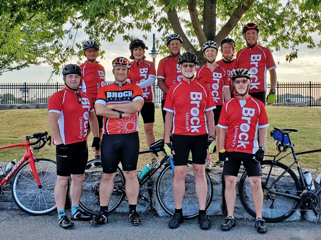  A team of 10 cyclists pose for a photograph. They are standing in two rows, all wearing red Brock University cycling jerseys. Three bicycles rest against a rock between the two rows. In the background is a tall tree and an iron fence.