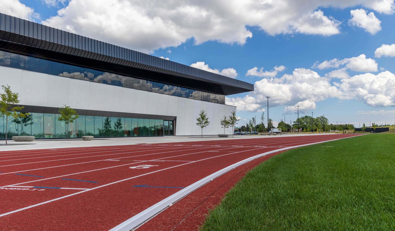 A red running track with green grass in the foreground and a modern sports facility in the background set against a blue sky with white clouds.