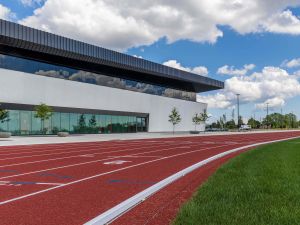 A red running track with green grass in the foreground and a modern sports facility in the background set against a blue sky with white clouds.