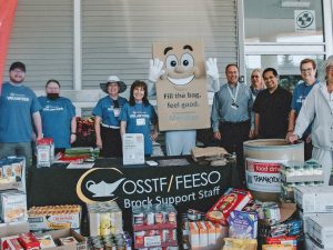 A group of people, joined by a mascot dressed as a paper bag, stand behind a table. In front of the table are stacks of non-perishable food items.