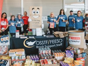A group of people, joined by a mascot dressed as a paper bag, stand behind a table. In front of the table are stacks of non-perishable food items.