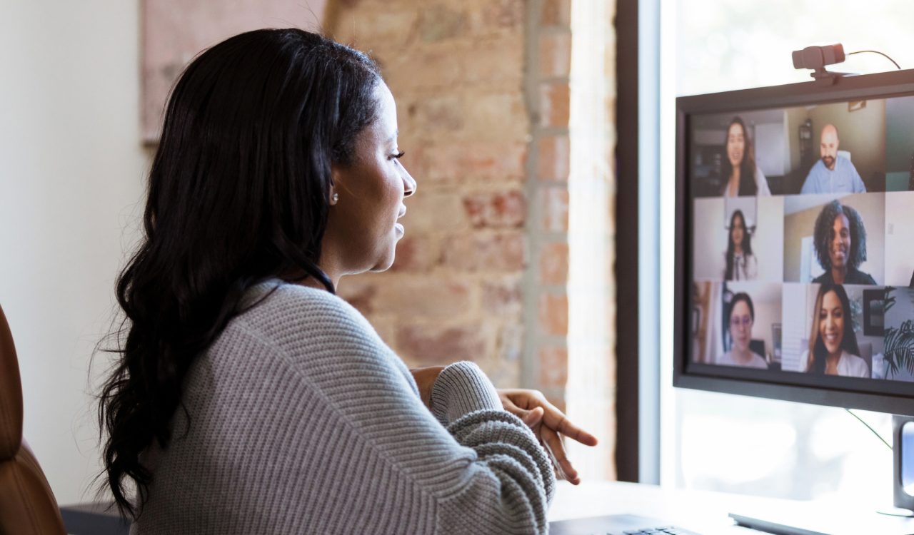 A woman attends an online meeting from home. She sits at a desk while looking at a computer screen that has a virtual meeting underway.