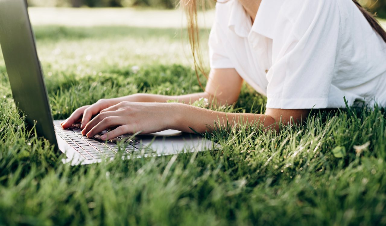A person with long hair and wearing a short-sleeved collared shirt is laying on their stomach in the grass and typing on a laptop.