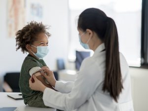 A young boy wearing a surgical mask has a bandaid placed on his arm by a female doctor.