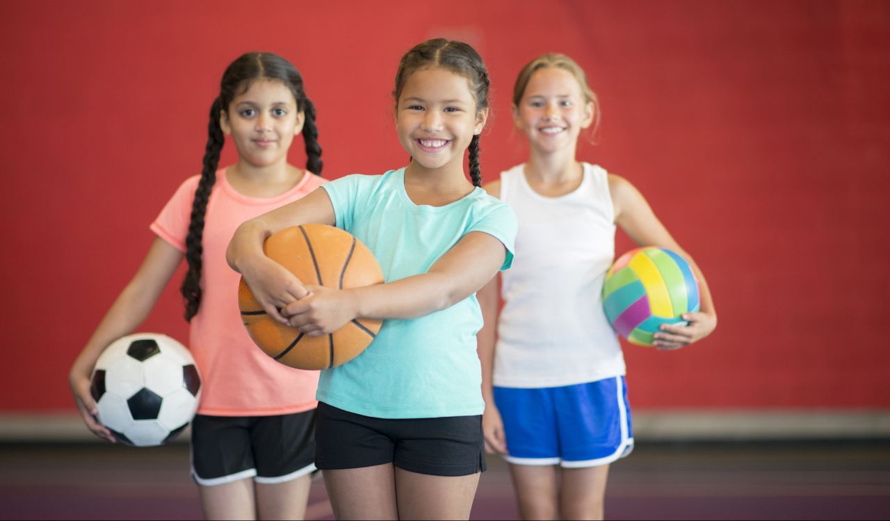 Three girls stand in front of a red wall in a gymnasium. The girl on the left is wearing a pink shirt and holding a soccer ball. The girl in the middle is wearing a teal shirt and holding a basketball. The girl on the right is wearing a white shirt and holding a volleyball.