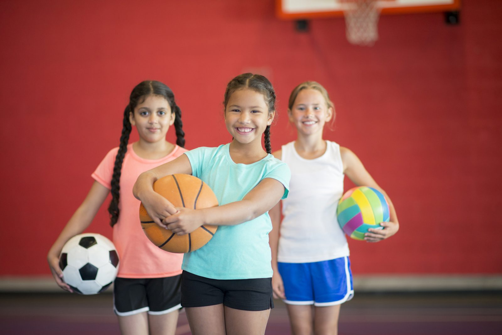 Three girls stand in front of a red wall in a gymnasium. The girl on the left is wearing a pink shirt and holding a soccer ball. The girl in the middle is wearing a teal shirt and holding a basketball. The girl on the right is wearing a white shirt and holding a volleyball.