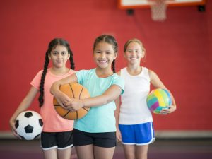 Three girls stand in front of a red wall in a gymnasium. The girl on the left is wearing a pink shirt and holding a soccer ball. The girl in the middle is wearing a teal shirt and holding a basketball. The girl on the right is wearing a white shirt and holding a volleyball.