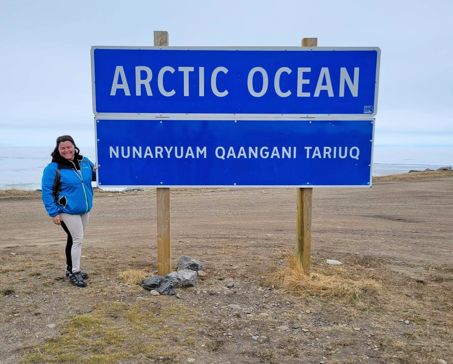 A woman in a blue jacket stands next to a blue sign that says ‘Arctic Ocean’ with the Arctic Ocean in the background.