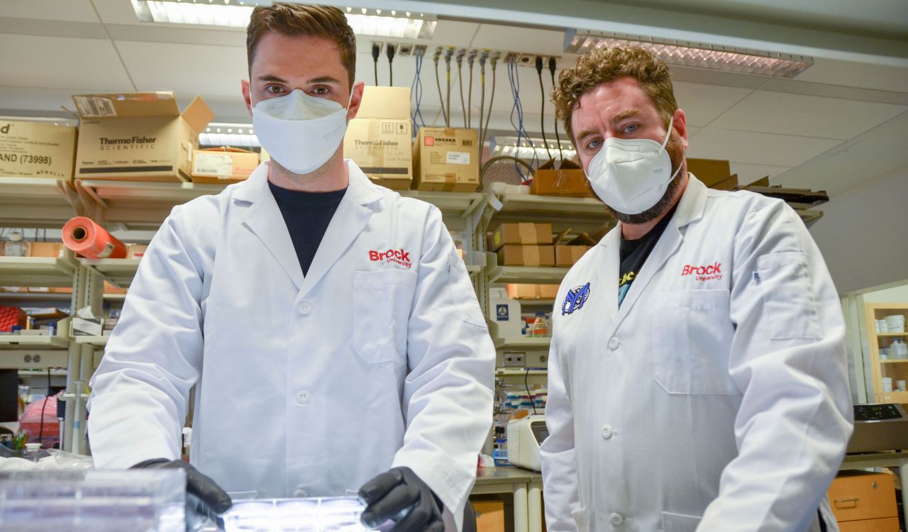 Waist-up photo of PhD student Jeremia Coish (left) and Associate Professor of Health Sciences Adam MacNeil (right) in the lab with boxes and shelves in the background. Both are wearing white Brock University lab coats, white masks and black shirts.
