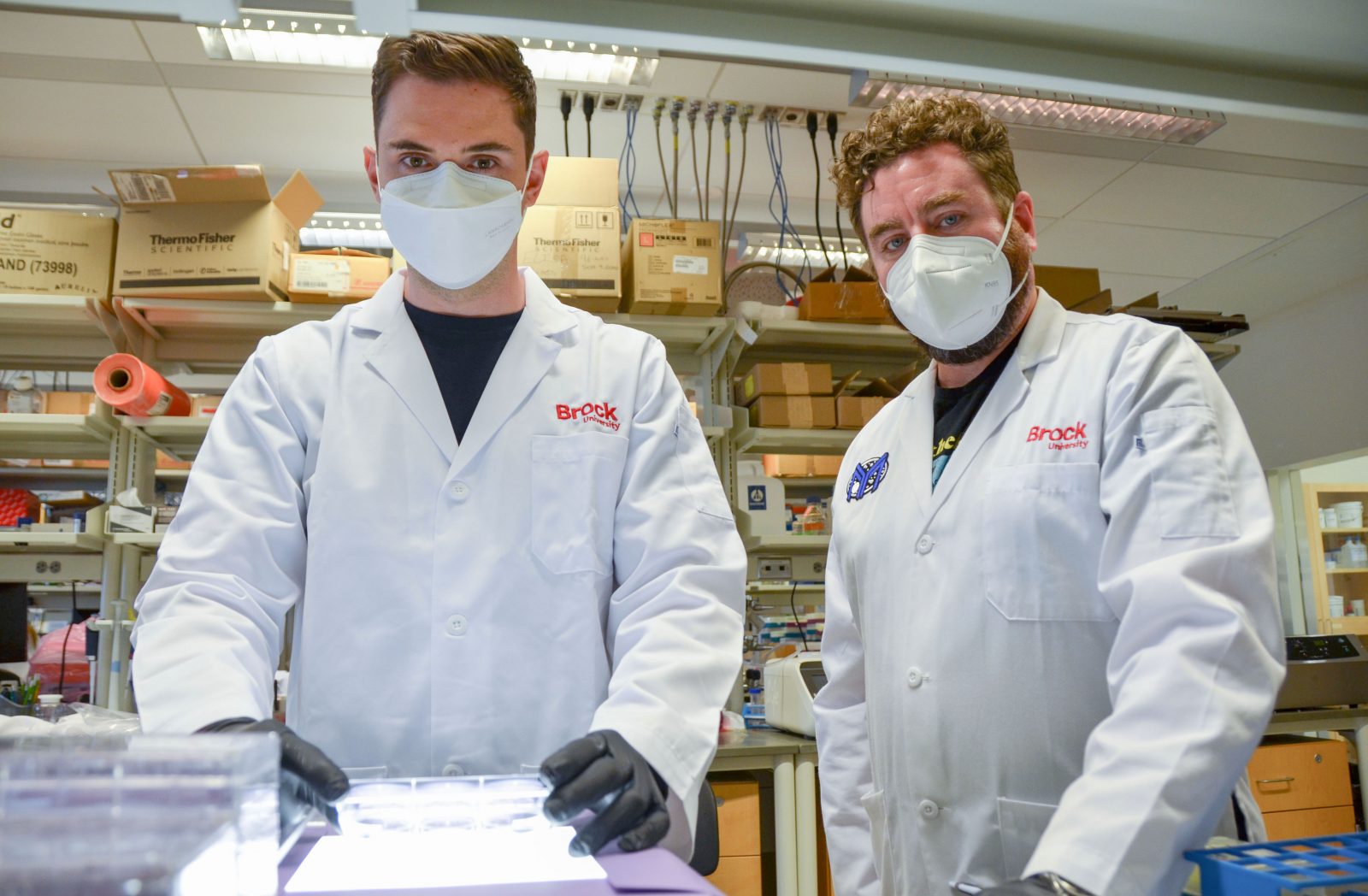 Waist-up photo of PhD student Jeremia Coish (left) and Associate Professor of Health Sciences Adam MacNeil (right) in the lab with boxes and shelves in the background. Both are wearing white Brock University lab coats, white masks and black shirts.