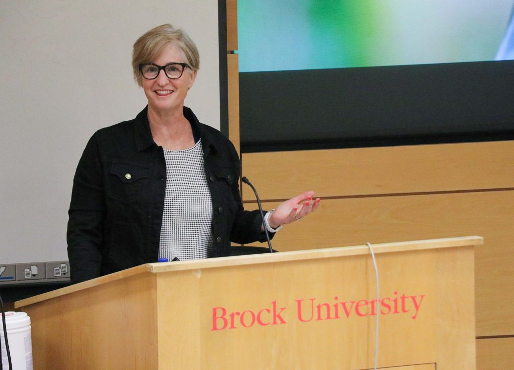 A woman with short blond hair and black thick-framed glasses stands at a podium that says 'Brock University.'