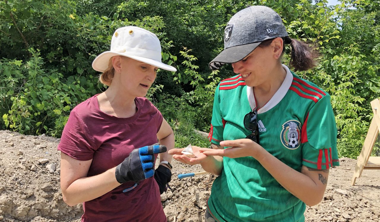 Two women stand outdoors discussing a small fragment of ceramic discovered during an archaeological dig.