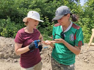 Two women stand outdoors discussing a small fragment of ceramic discovered during an archaeological dig.