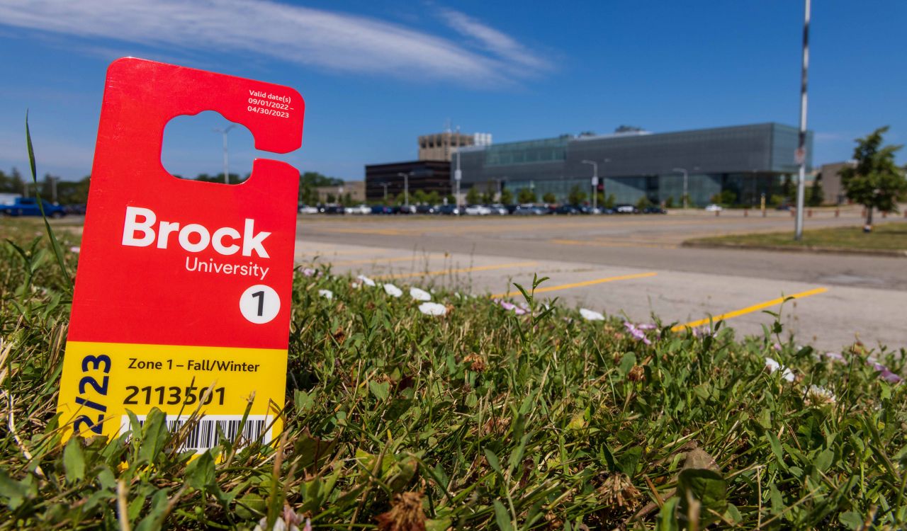 A red parking permit with the work 'Brock' in white letters sits in a patch of grass with a parking lot and glass building in the background.