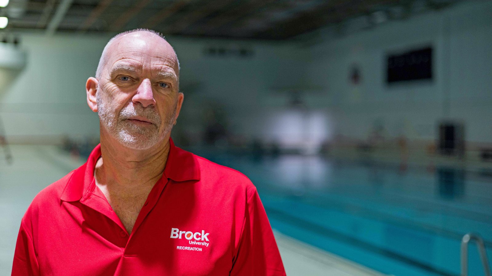 A man in a red Brock University shirt stands beside a pool.