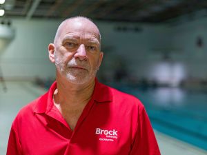 A man in a red Brock University shirt stands beside a pool.
