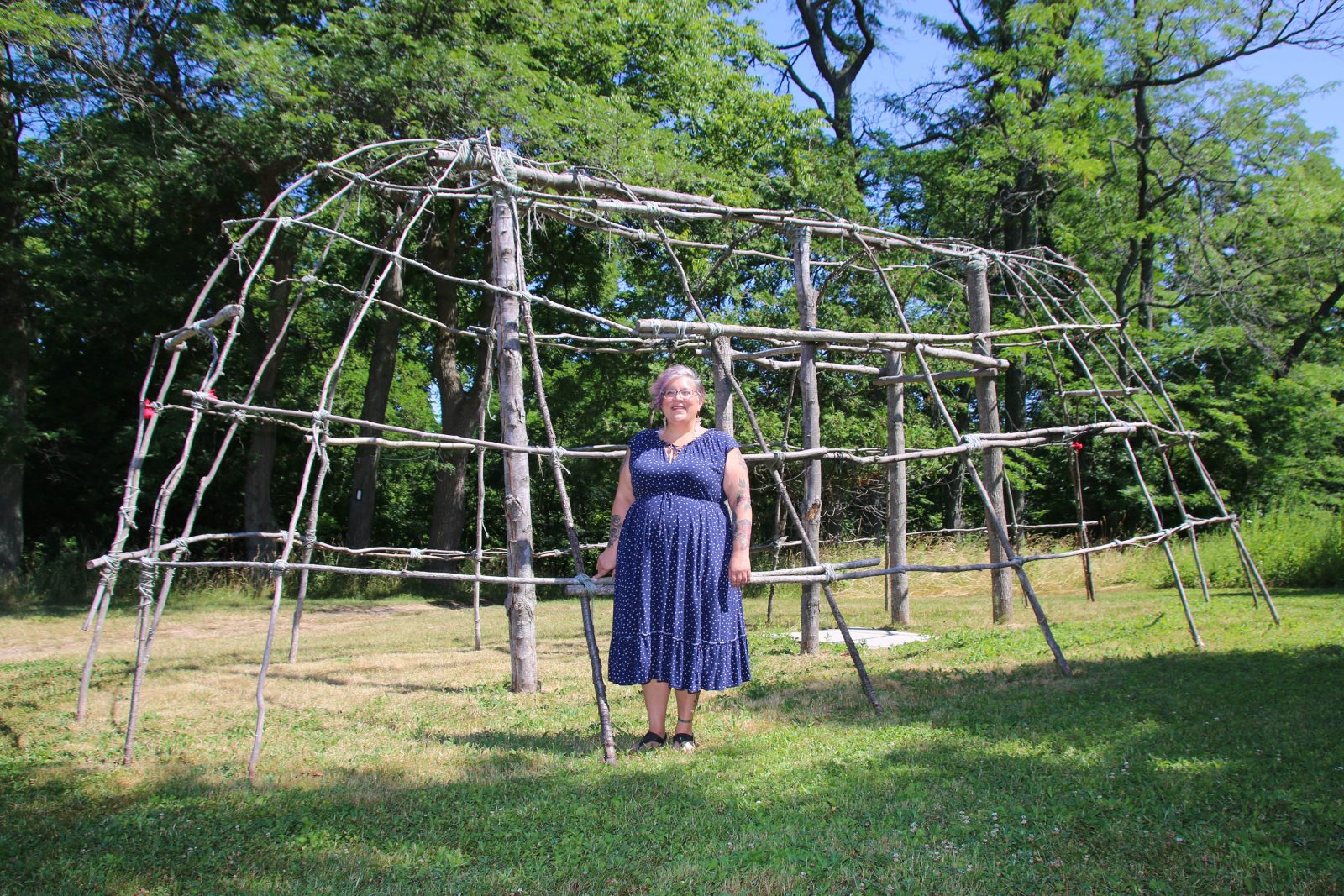A woman in a blue dress stands in a field of grass in front of an oval-shaped building frame made from wooden branches.