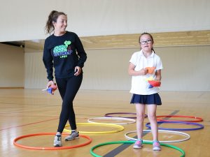 A young woman and a young girl stand together in a gymnasium with coloured hula hoops on the floor. They each hold colourful bean bags in their hands.