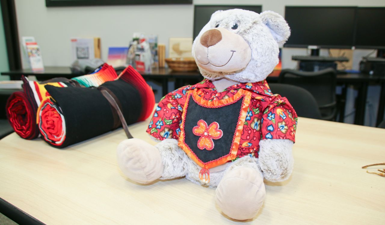 A stuffed bear clothed in a red and blue printed shirt and an orange and black sash sits on a table.