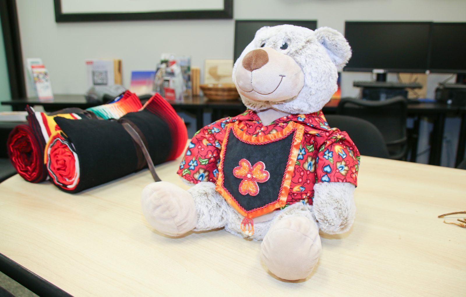 A stuffed bear clothed in a red and blue printed shirt and an orange and black sash sits on a table.