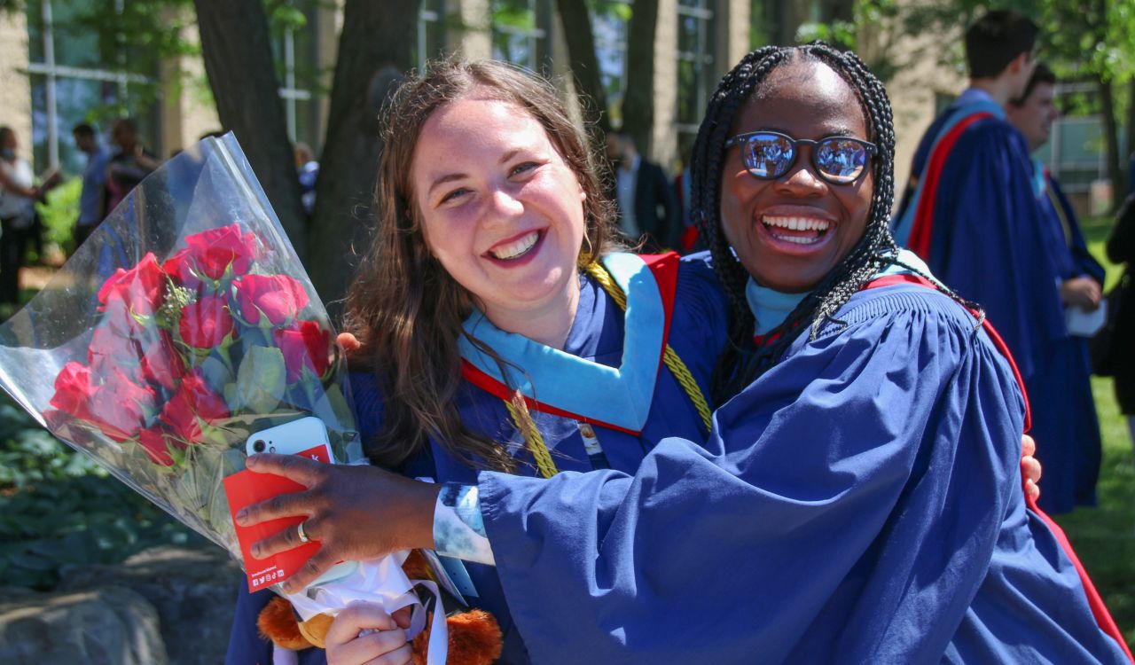 Two women in blue graduation gowns hug one another, one holding a bouquet of flowers.