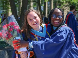 Two women in blue graduation gowns hug one another, one holding a bouquet of flowers.