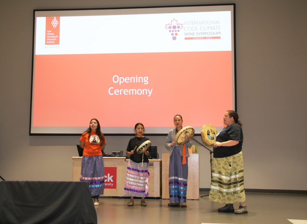 Four women are pictured singing while playing traditional drums in front of a large red screen that says 'opening ceremony.'