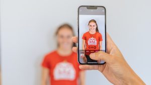 A woman has her photo taken with a smartphone while standing against a white wall.