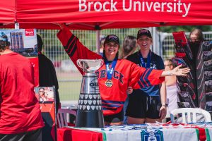 Two female student-athletes in Brock Badger fan gear pose under a red tent with a silver trophy.