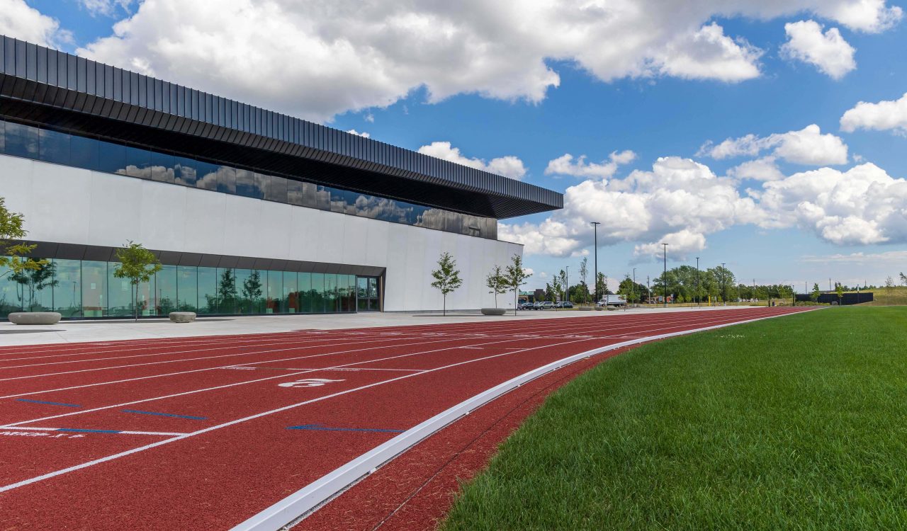 A red running track with green grass in the foreground and a modern sports facility in the background set against a blue sky with white clouds.