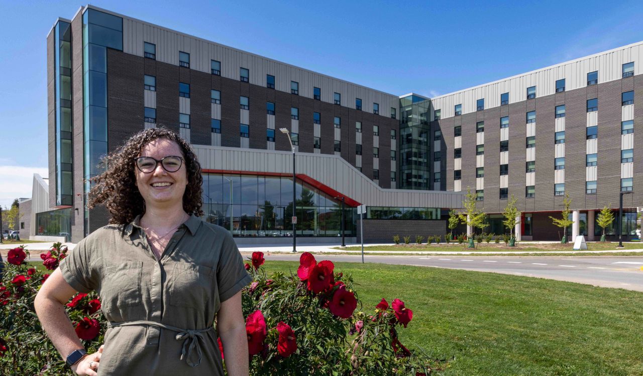 A woman with curly brown hair and glasses stands in front of a large patch of red hibiscus flowers. In the background is a large, six-floor university residence building with grey brick and many windows.