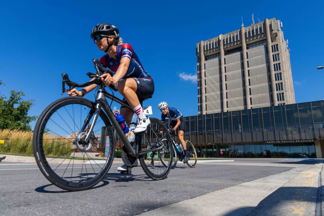 A closeup of two cyclists riding on a roadway in front of a large concrete tower.