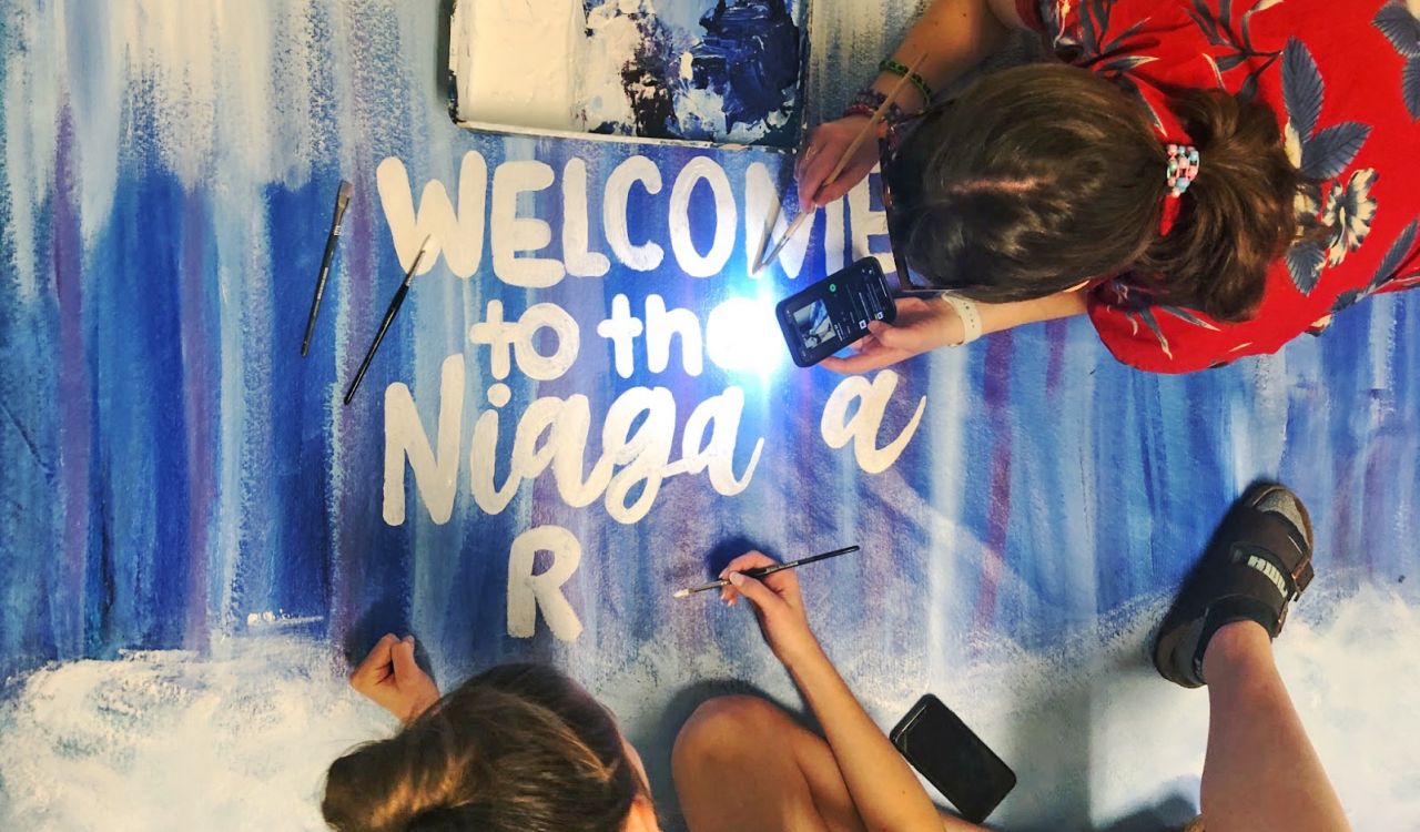 A photo taken from above shows two young girls in progress of painting a canvas that will read 'Welcome to the Niagara region.'