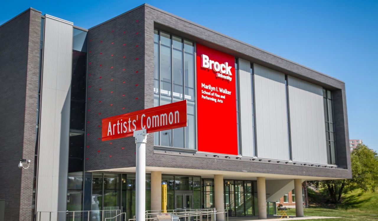 A grey and red building sits against a clear, blue sky with green grass surrounding it. The building has white text on it on a red background reading ‘Marilyn I. Walker School of Fine and Performing Arts, Brock University.’ A street sign reading ‘Artists’ Common’ sits in the foreground.