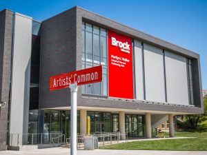A grey and red building sits against a clear, blue sky with green grass surrounding it. The building has white text on it on a red background reading ‘Marilyn I. Walker School of Fine and Performing Arts, Brock University.’ A street sign reading ‘Artists’ Common’ sits in the foreground.