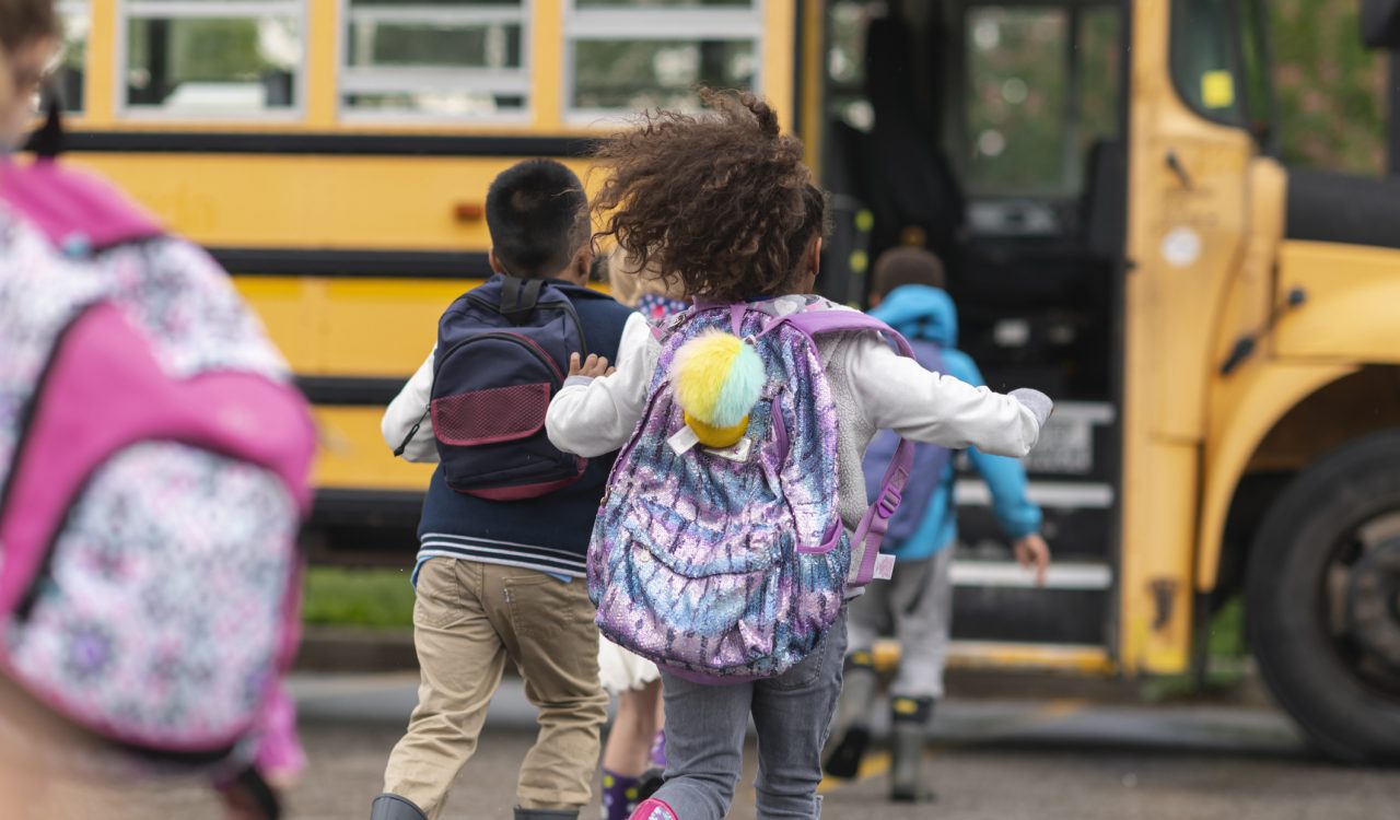 Children wearing backpacks run towards a yellow school bus.