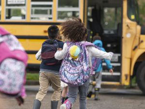 Children wearing backpacks run towards a yellow school bus.
