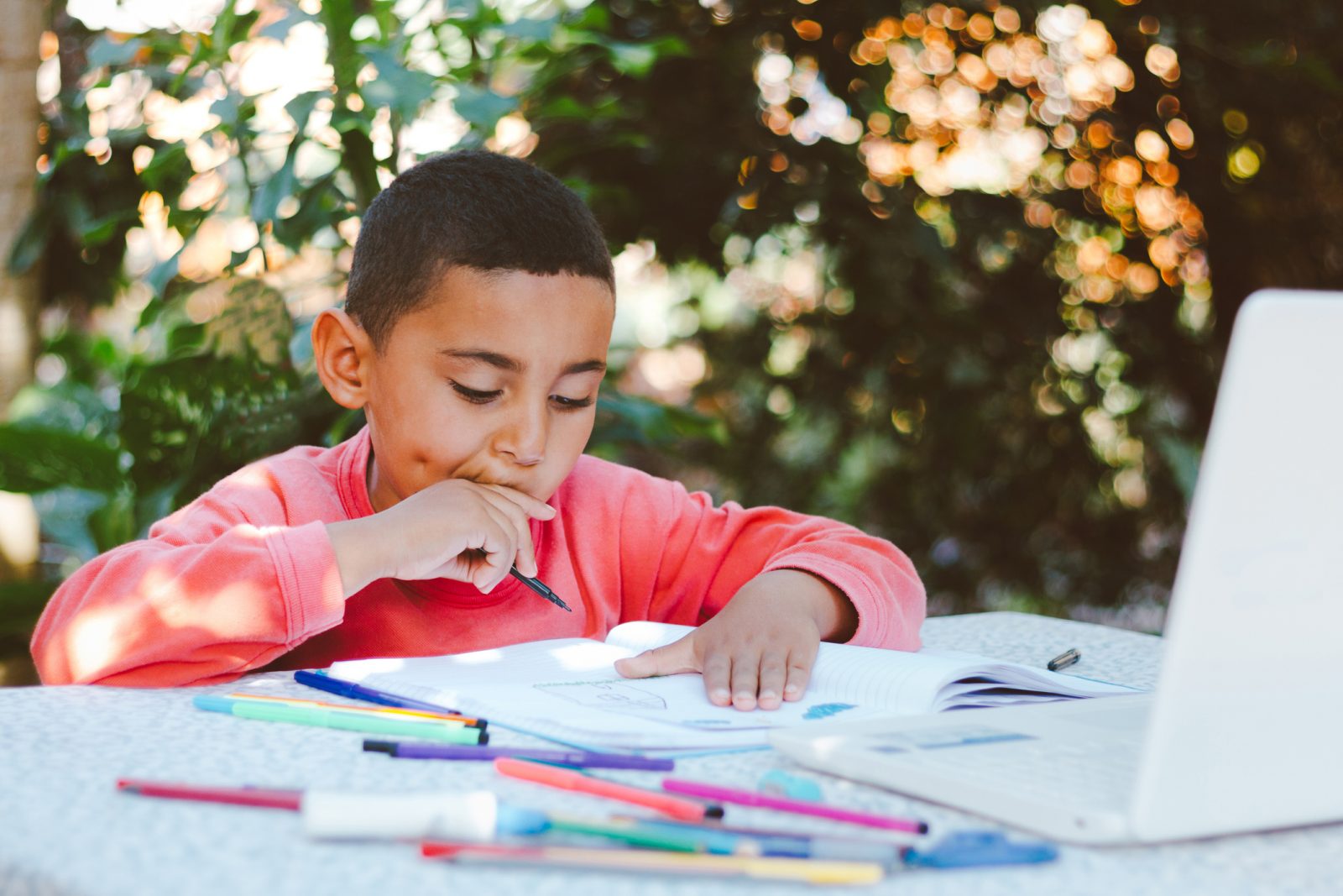 A boy sits at a table outdoors writing and drawing in a notebook with colourful markers.