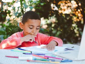 A boy sits at a table outdoors writing and drawing in a notebook with colourful markers.