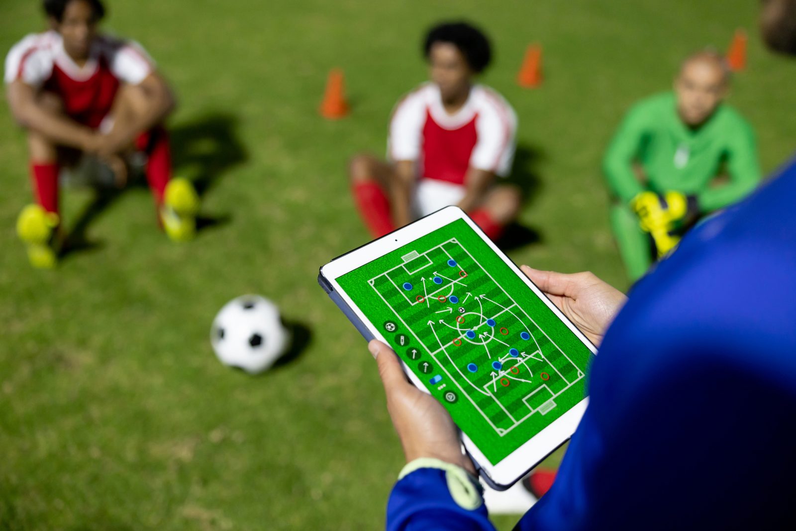 A tablet in the foreground being held by two hands shows a soccer field with arrows, lines and dots, with three soccer players on a green field are sitting with a soccer ball in a blurred background.