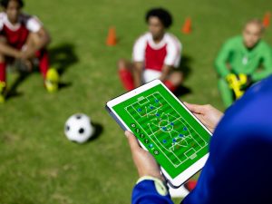 A tablet in the foreground being held by two hands shows a soccer field with arrows, lines and dots, with three soccer players on a green field are sitting with a soccer ball in a blurred background.