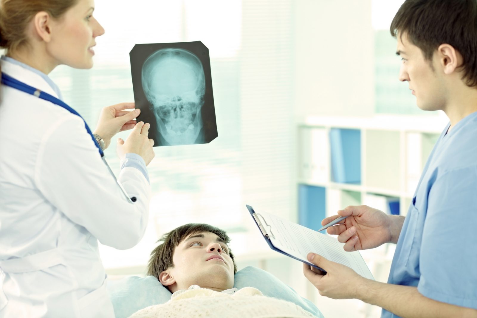 A man with brown hair is lying on a hospital bed looking up at a female medical health professional (left) holding an X-ray of a skull with a male medical professional (right) looking at the X-ray and holding a clipboard and pen.