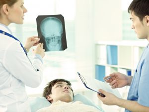A man with brown hair is lying on a hospital bed looking up at a female medical health professional (left) holding an X-ray of a skull with a male medical professional (right) looking at the X-ray and holding a clipboard and pen.