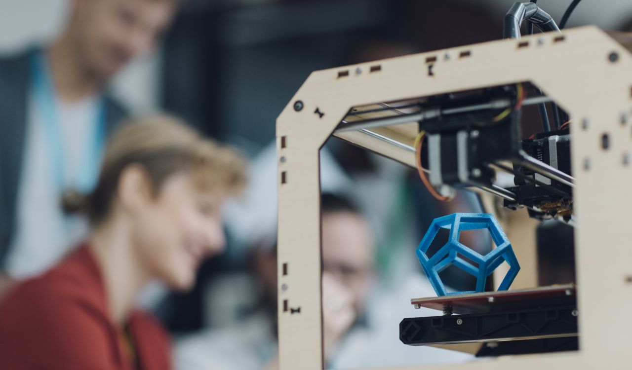 A 3D printer carves a geometric object while two blurred figures work in the background.