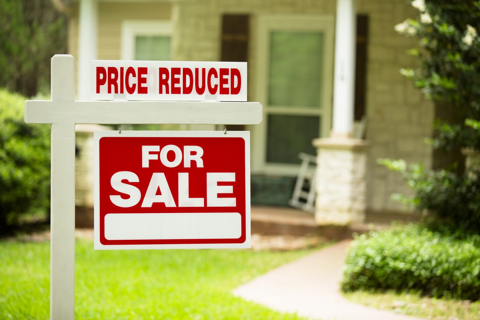 Close-up of a red-and-white sign saying ‘Price reduced, For Sale’ with a blurred white porch and green trees in the background.
