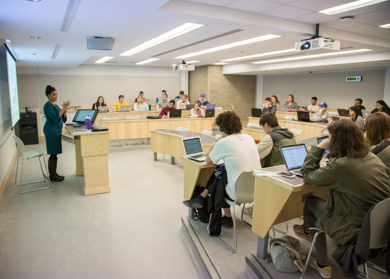 Woman stands at podium with a computer screen in small classroom. Rows of students sit at long, curved tables facing her.