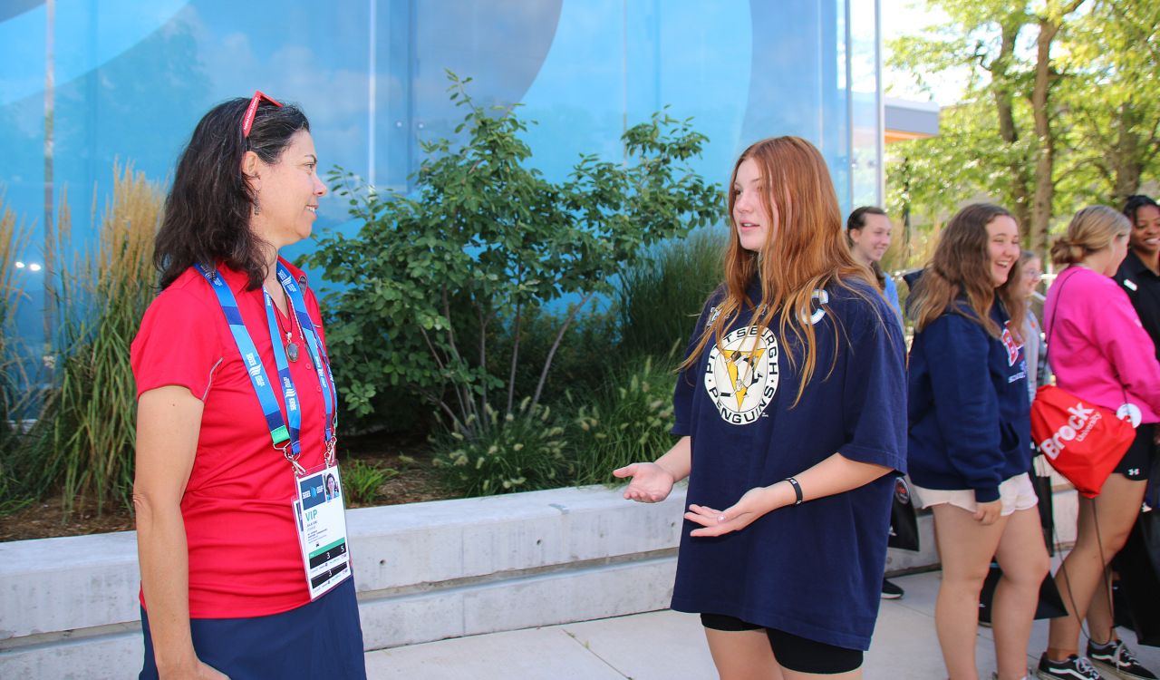 A woman in a red shirt speaks to a girl in a blue shirt outdoors in front of a blue background.