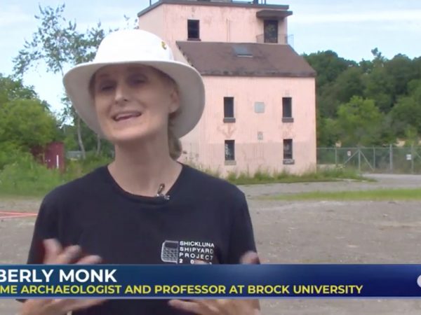 A woman in a blue shirt and white hat stands in front of an open gravel lot and a pink building.
