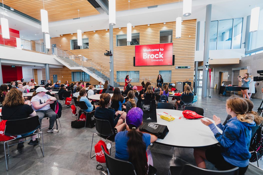 An atrium filled with people who sit at a series of round tables and look up at the front where a screen reads 'Welcome to Brock University.'